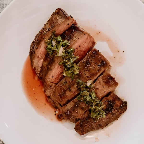 Sliced steak with a pink center, topped with green herbs and resting in its juices on a white plate.