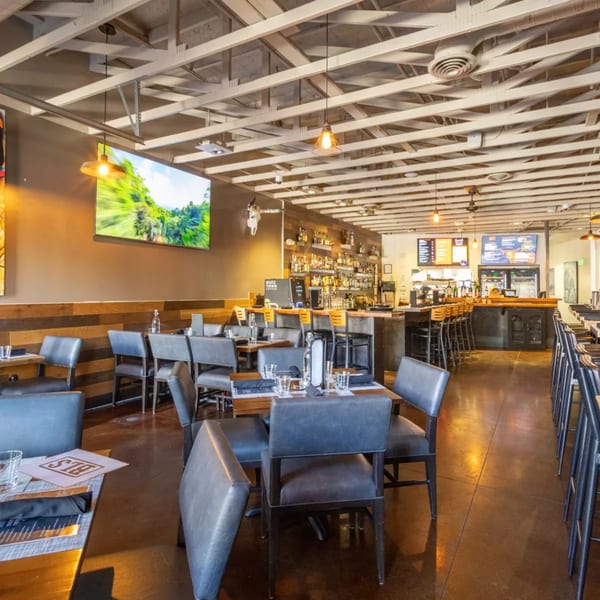 Modern restaurant interior with dark tables, gray chairs, a large screen, and a well-stocked bar.