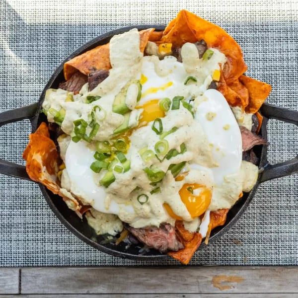 Skillet of chilaquiles with steak, fried eggs, creamy sauce, avocado, and green onions.