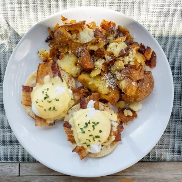 Eggs Benedict with bacon and hollandaise sauce, served with crispy smashed potatoes on a white plate.