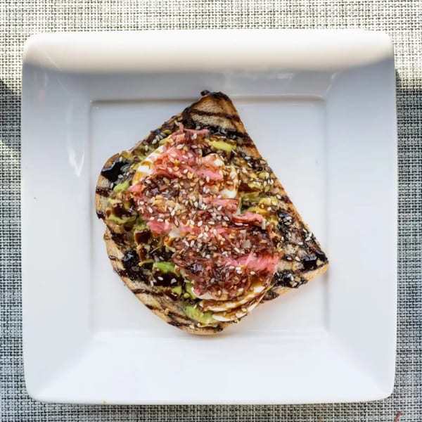 Grilled toast topped with avocado, pink pickled onions, white sauce, and sesame seeds.