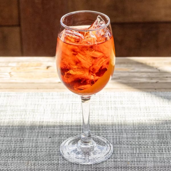 A vibrant orange cocktail with ice and an orange slice in a wine glass on a textured grey surface.
