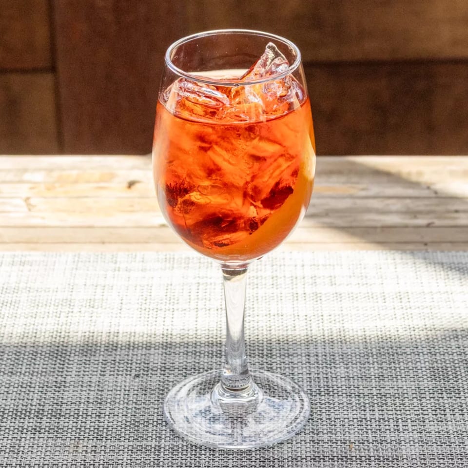 A vibrant orange cocktail with ice and an orange slice in a wine glass on a textured grey surface.