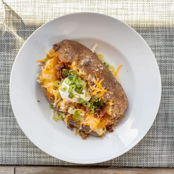 A loaded baked potato with shredded cheese, bacon bits, sour cream, and green onions on a white plate.
