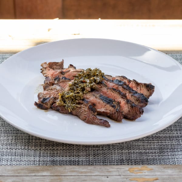 Grilled skirt steak slices topped with green chimichurri sauce on a white plate.