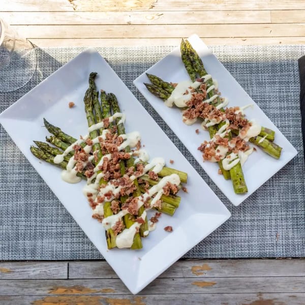 Two plates of grilled asparagus drizzled with white sauce and topped with brown crumbles on a wooden table.